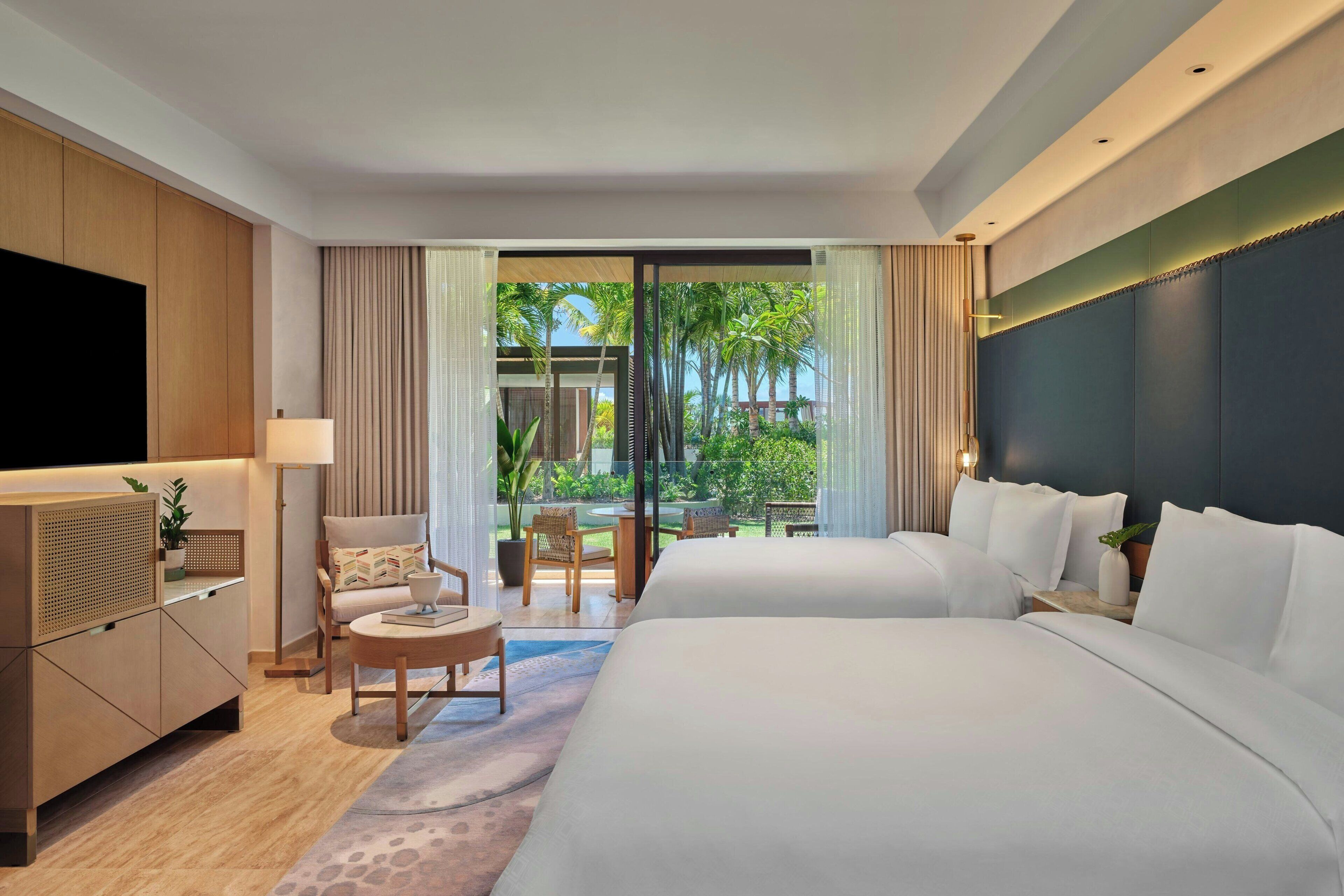 A hotel room with two beds, a TV, a desk, and a view of a garden.