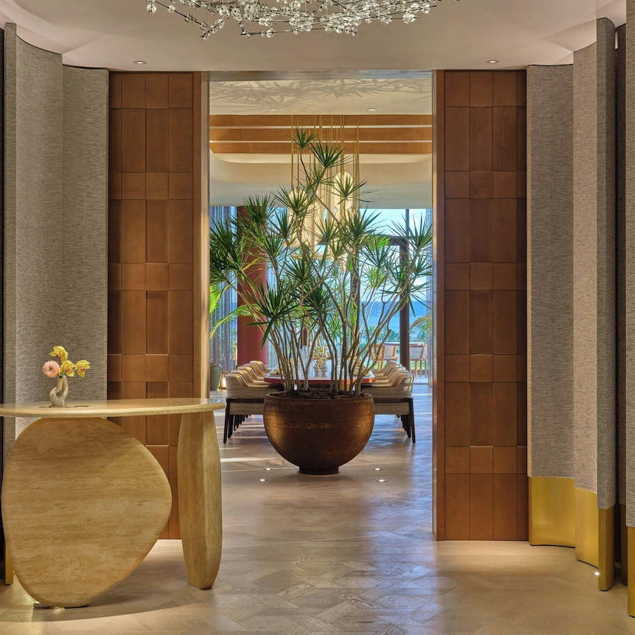 A modern hotel lobby with a curved wooden reception desk, a large potted plant, and a seating area with chairs.