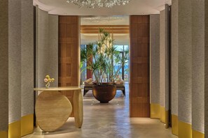 A modern hotel lobby with a curved wooden reception desk, a large potted plant, and a seating area with chairs.