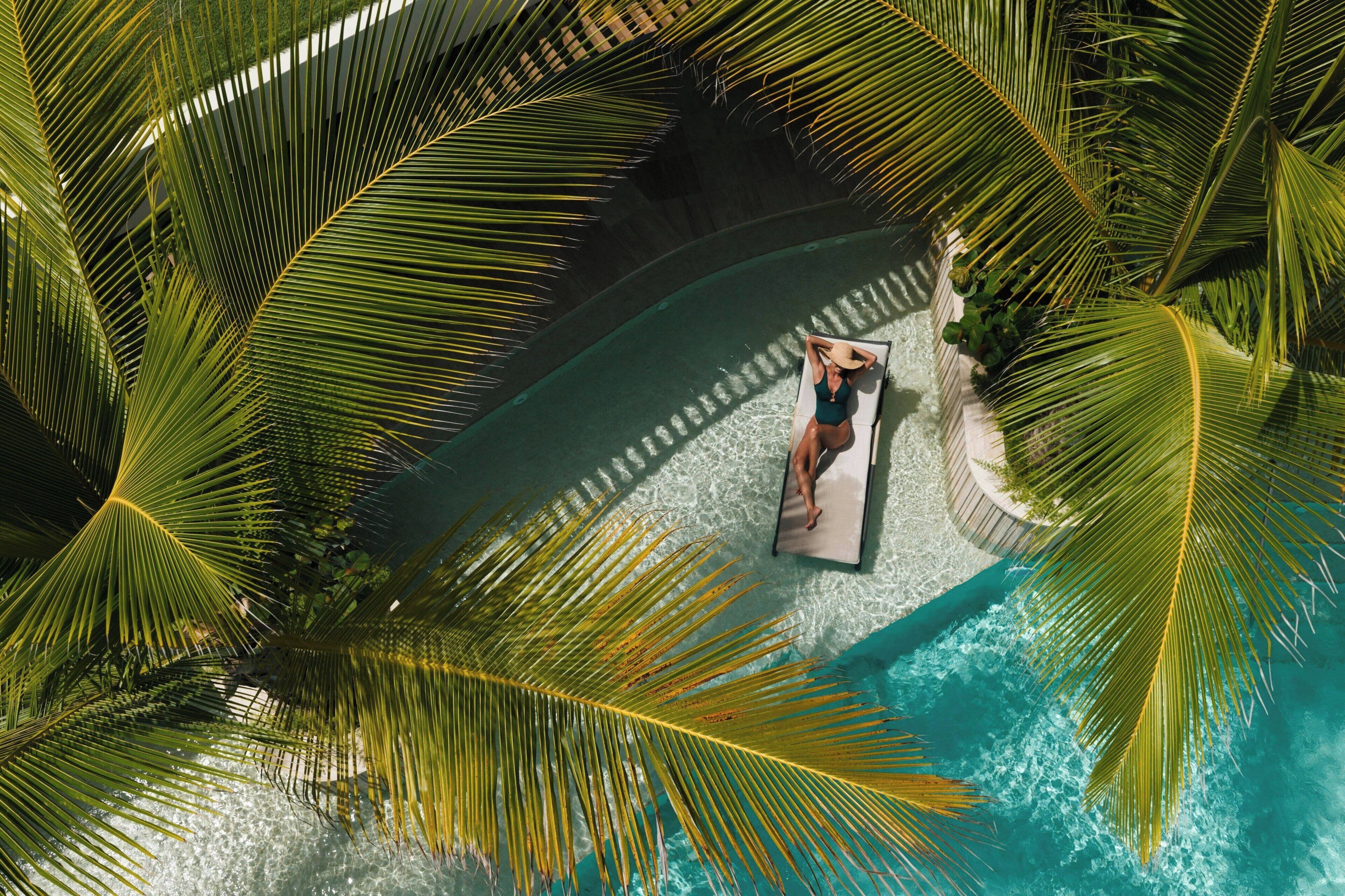A person relaxing on a floating platform in a pool surrounded by palm trees.