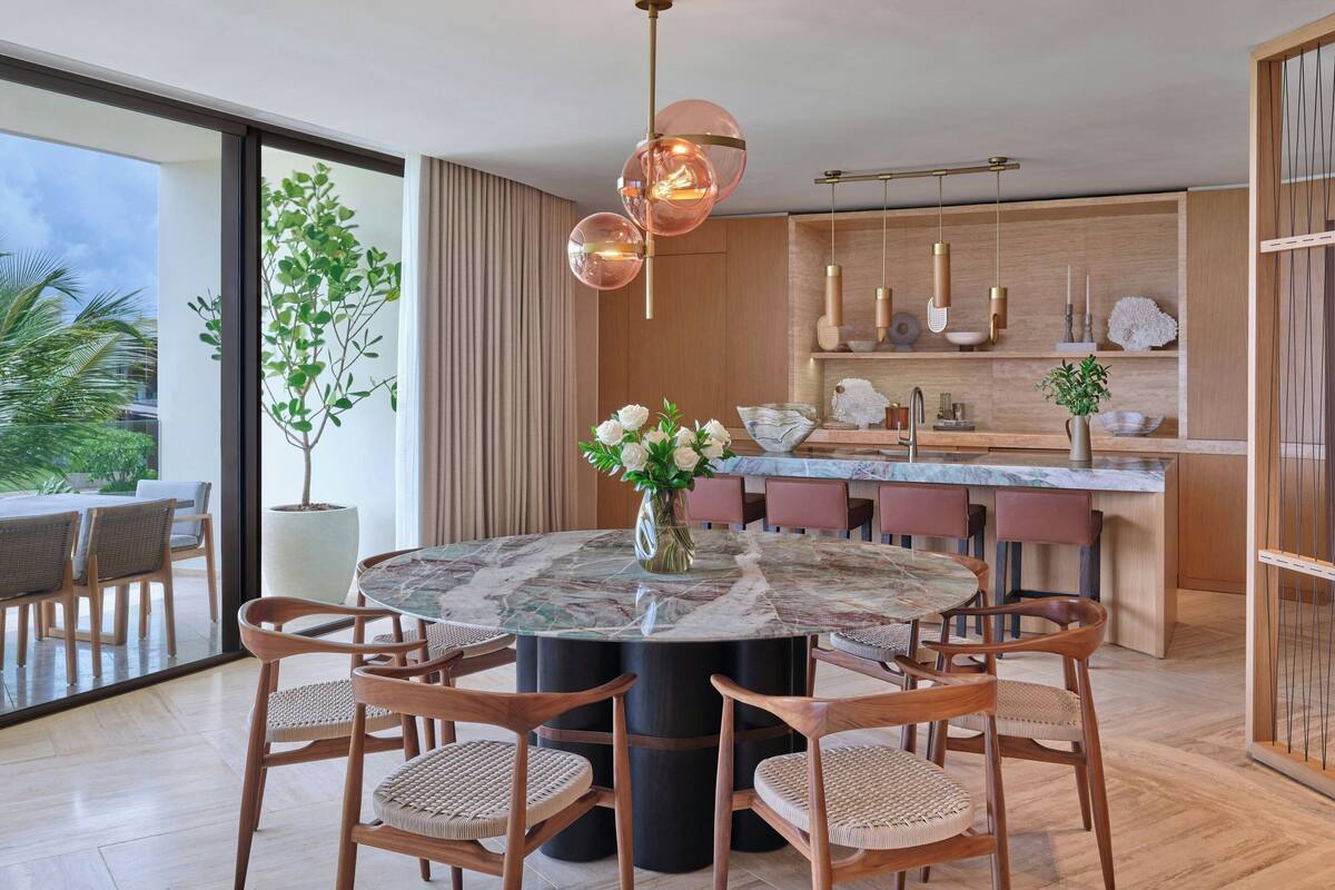 A modern dining area with a round table, six chairs, a vase of flowers, and a large potted plant.