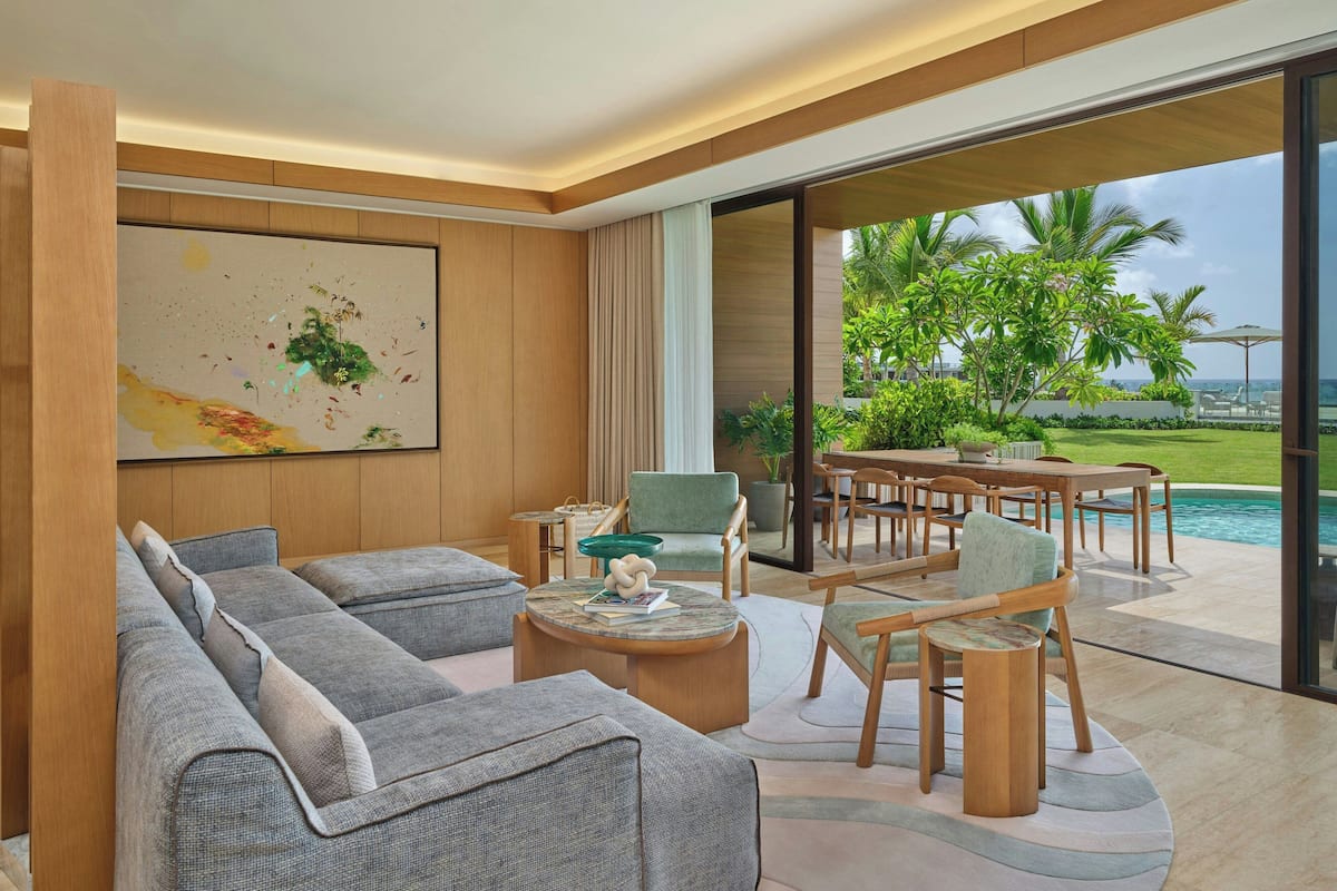 A modern living room with a large sofa, a coffee table, and a dining area with chairs. There is a large window with a view of a pool and palm trees.