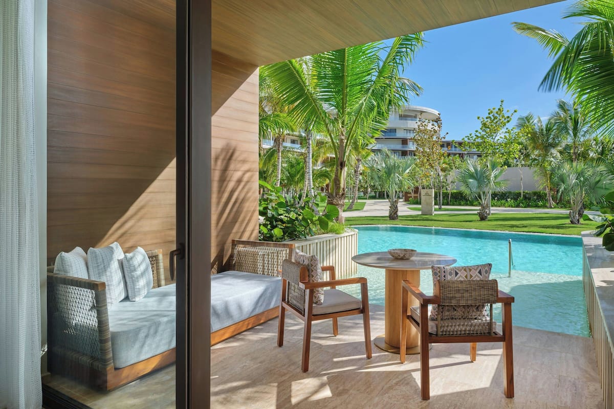 A balcony with a bed, two wicker chairs, a small table, and a view of a swimming pool and palm trees.