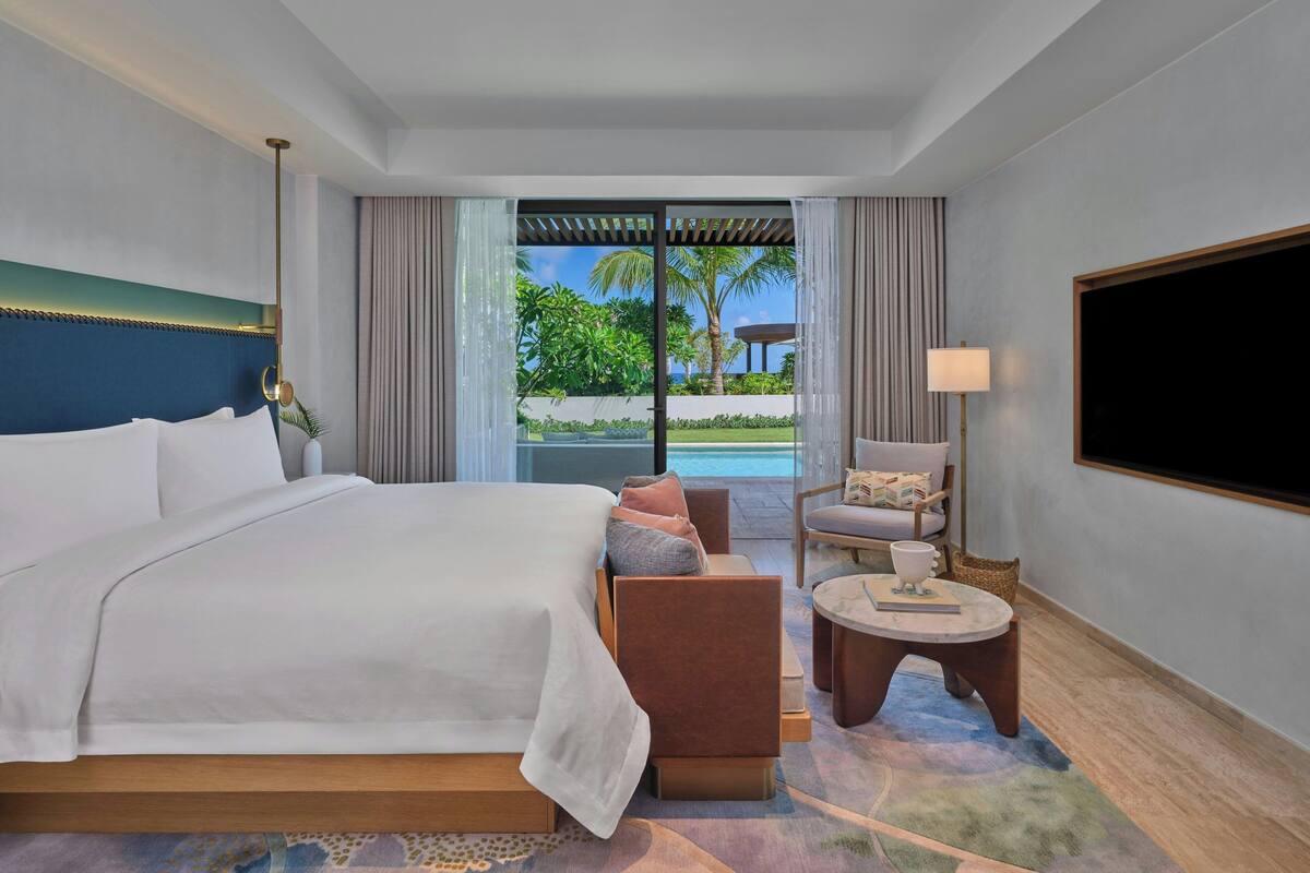 A hotel room with a bed, a seating area with a table, a TV, and a view of a pool and greenery.