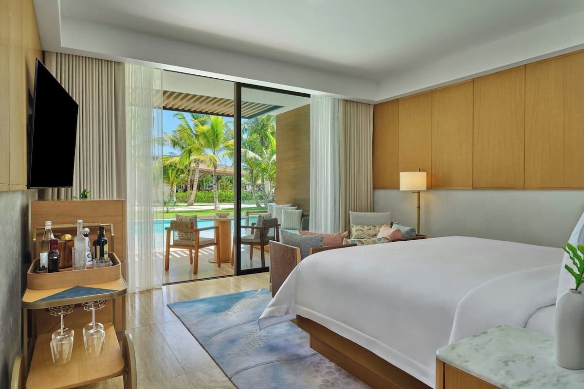 A modern hotel room with a large bed, a TV, a minibar, and a view of a pool and palm trees.