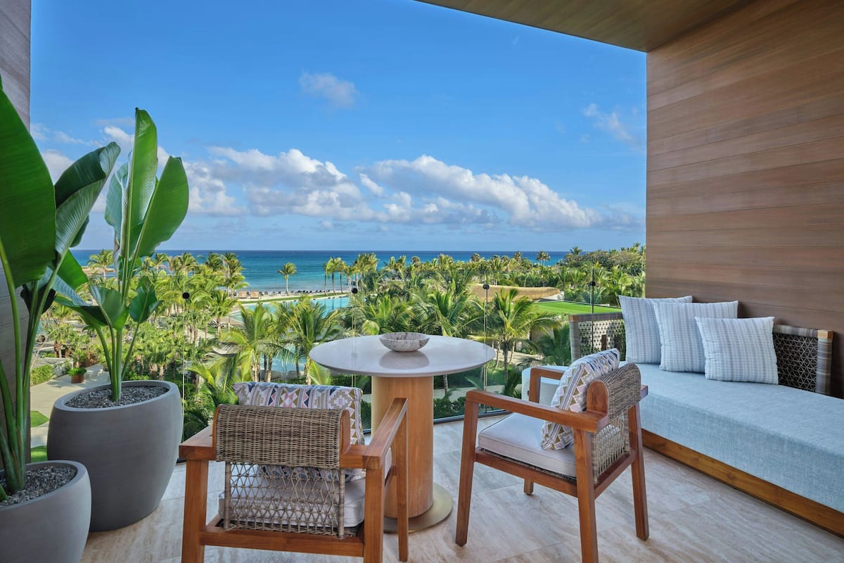 A balcony with a view of the ocean, a round table, two wicker chairs, a sofa with cushions, and large potted plants.