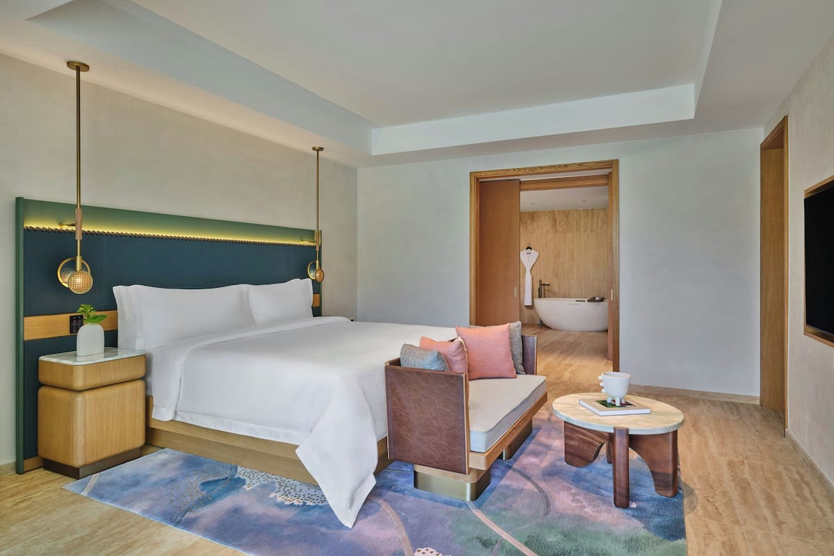 A modern hotel room with a large bed, bedside tables, a small sofa, a round coffee table, and a bathroom visible through an open door.