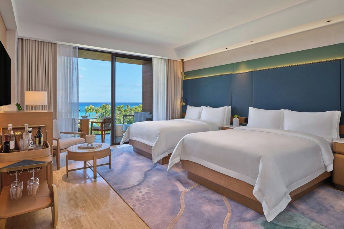 A hotel room with two beds, a desk with a lamp, a chair, a small table, and a view of the ocean.
