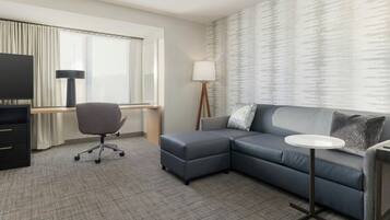 A modern hotel room with a grey sectional sofa, a small round table, a desk with a chair, a TV, and a large window with curtains.