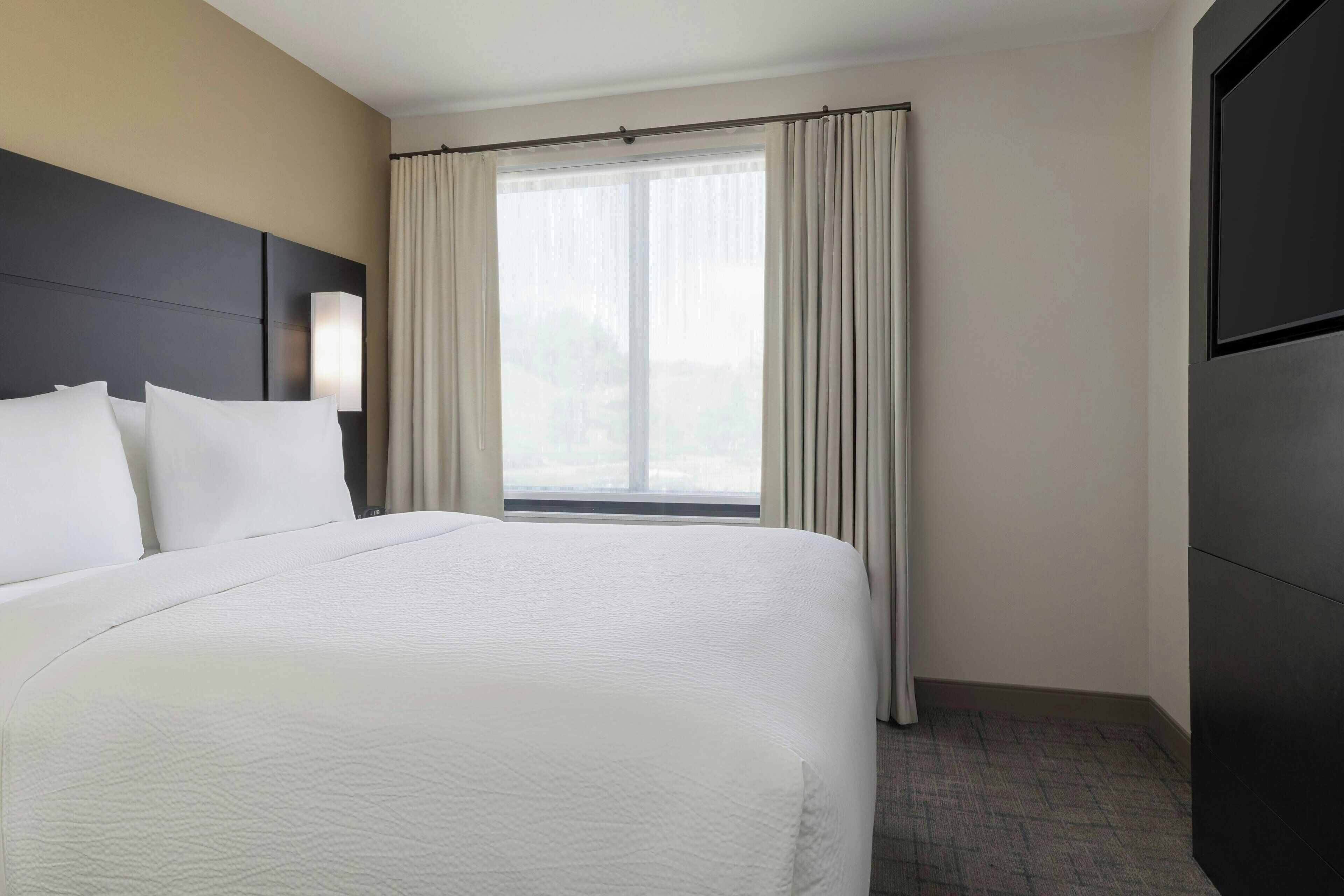 Habitación de hotel con una cama grande, cabecero, ventana con cortinas y televisor montado en la pared.