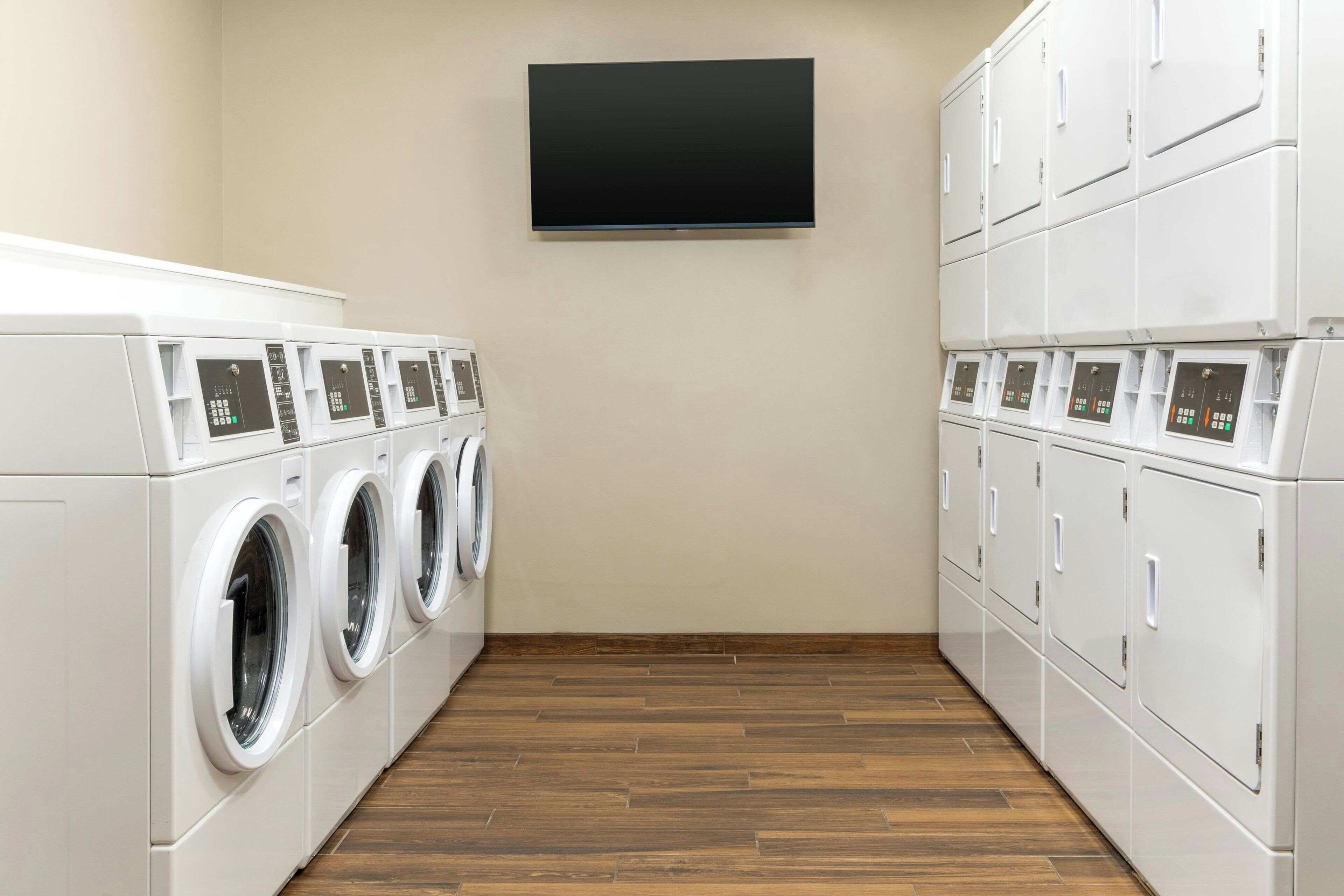 A laundry room with multiple washing machines and dryers, a flat-screen TV mounted on the wall, and wooden flooring.
