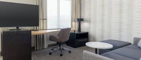 A hotel room with a desk, chair, sofa, TV, lamp, and a window with curtains.