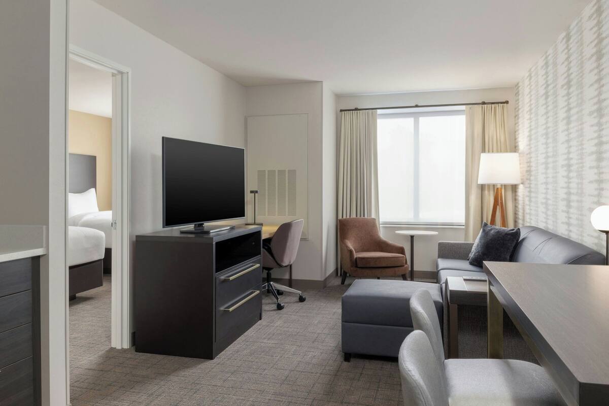 A hotel room with a bed, a TV, a desk, a chair, a sofa, and a window with curtains.