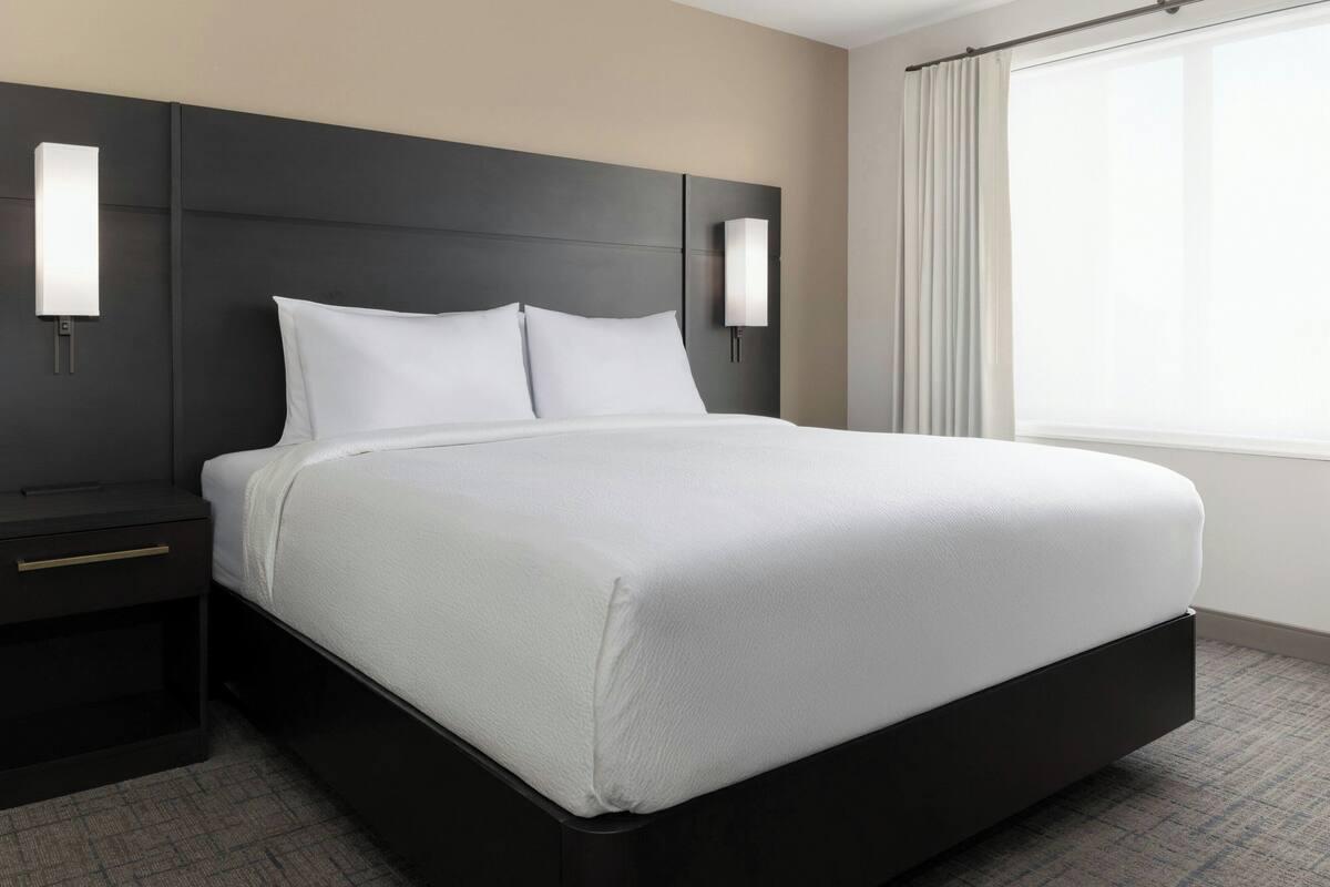 A modern hotel room with a large bed, dark headboard, bedside tables, and wall-mounted light fixtures.