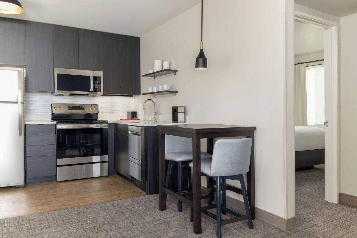 A compact kitchen with modern appliances, a dining area with bar stools, and a visible bedroom in the background.