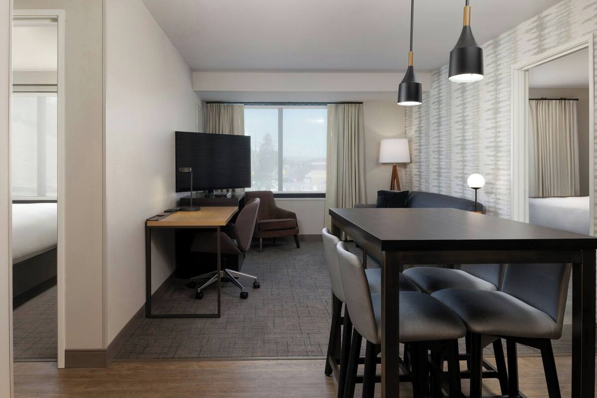 A hotel room with a desk, chairs, a television, and a large window with curtains.