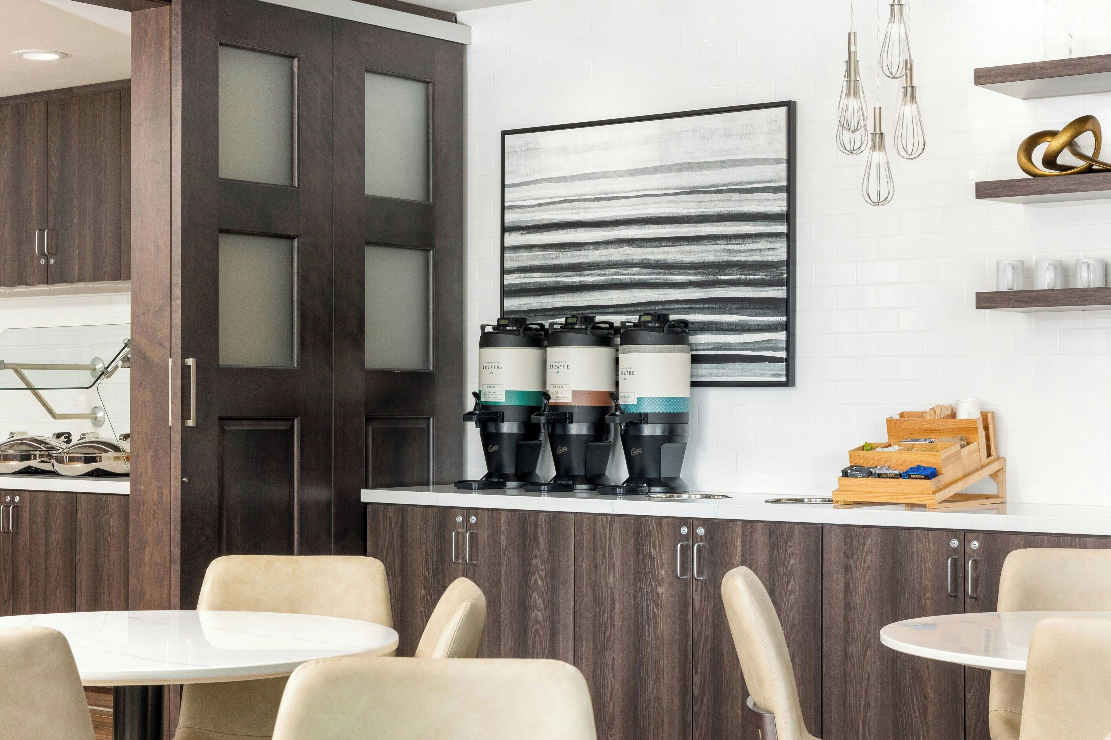 A modern kitchen with dark wood cabinets, a white countertop, and a coffee station with multiple coffee makers. There are two round tables with chairs, and a large abstract painting on the wall.