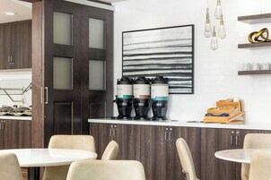 A modern kitchen with dark wood cabinets, a white countertop, and a coffee station with multiple coffee makers. There are two round tables with chairs, and a large abstract painting on the wall.