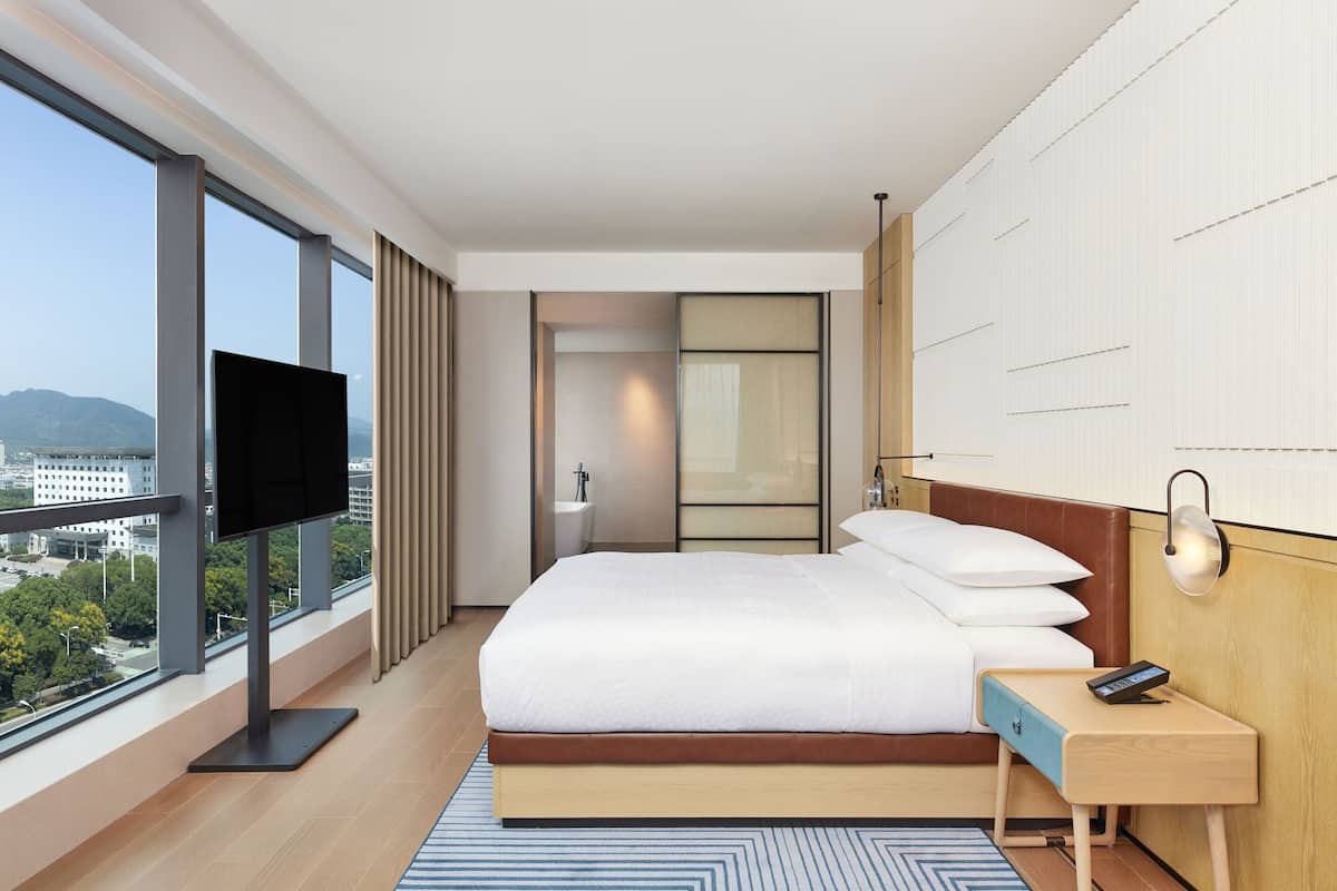 Una habitación de hotel moderna con una cama grande, televisión, mesita de noche y vista a la ciudad.