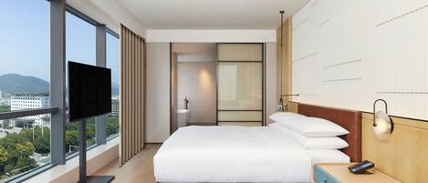 A modern hotel room with a large bed, a TV, a bedside table, and a view of the city.