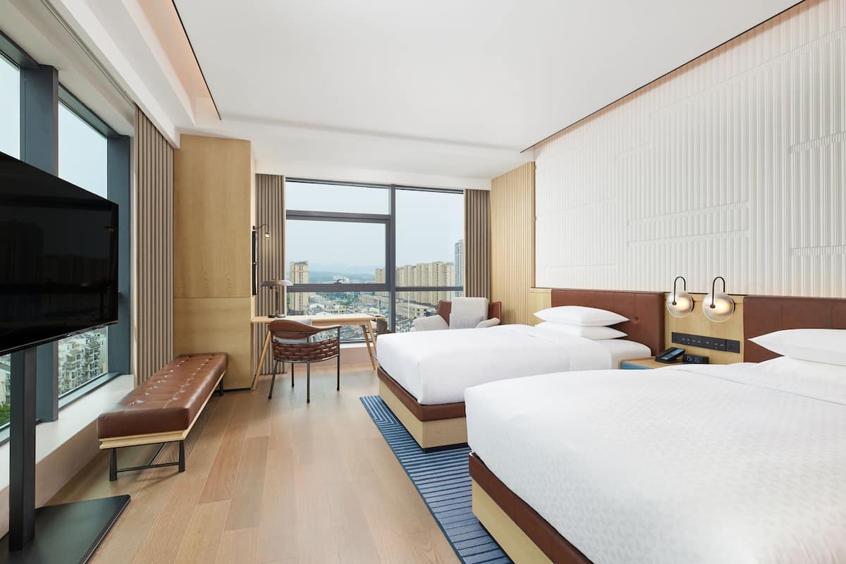 Habitación de hotel con dos camas, televisión, escritorio y un ventanal con vista a la ciudad.