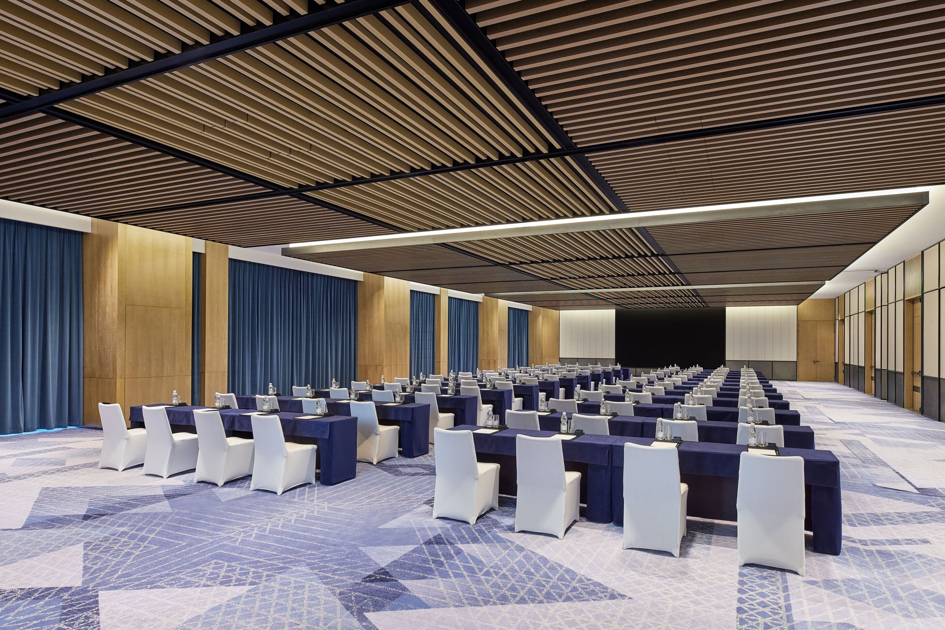 A conference room with rows of tables and chairs, a patterned carpet, and wooden paneling on the ceiling.