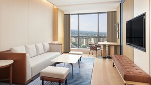 Una habitación de hotel moderna con un ventanal grande, un sofá, una mesa de centro, un banco y un televisor.