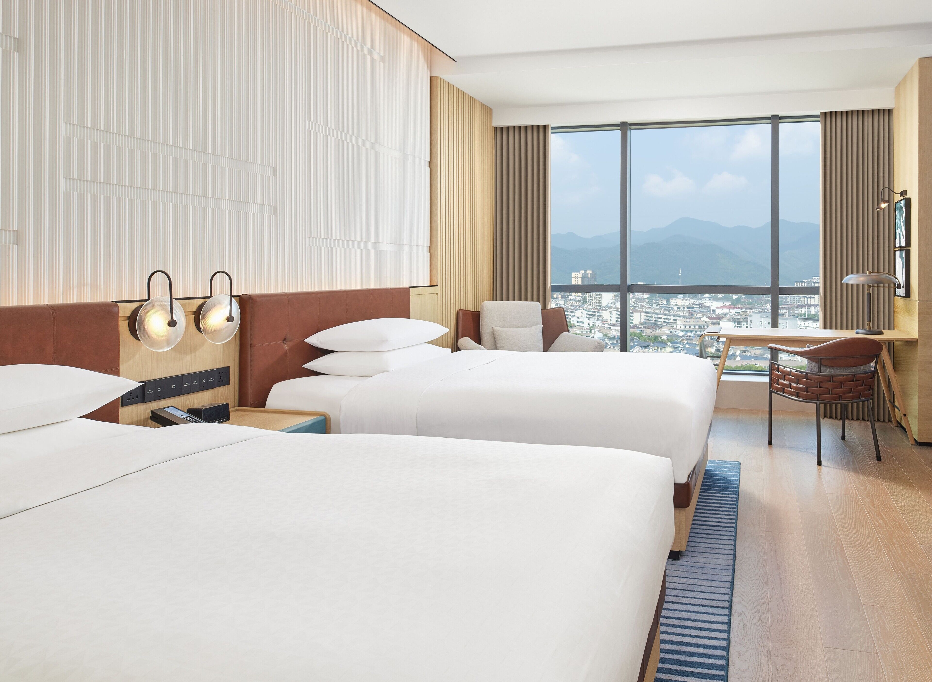 Une chambre d’hôtel avec deux lits, un bureau, une chaise et une grande fenêtre donnant sur la ville et les montagnes.
