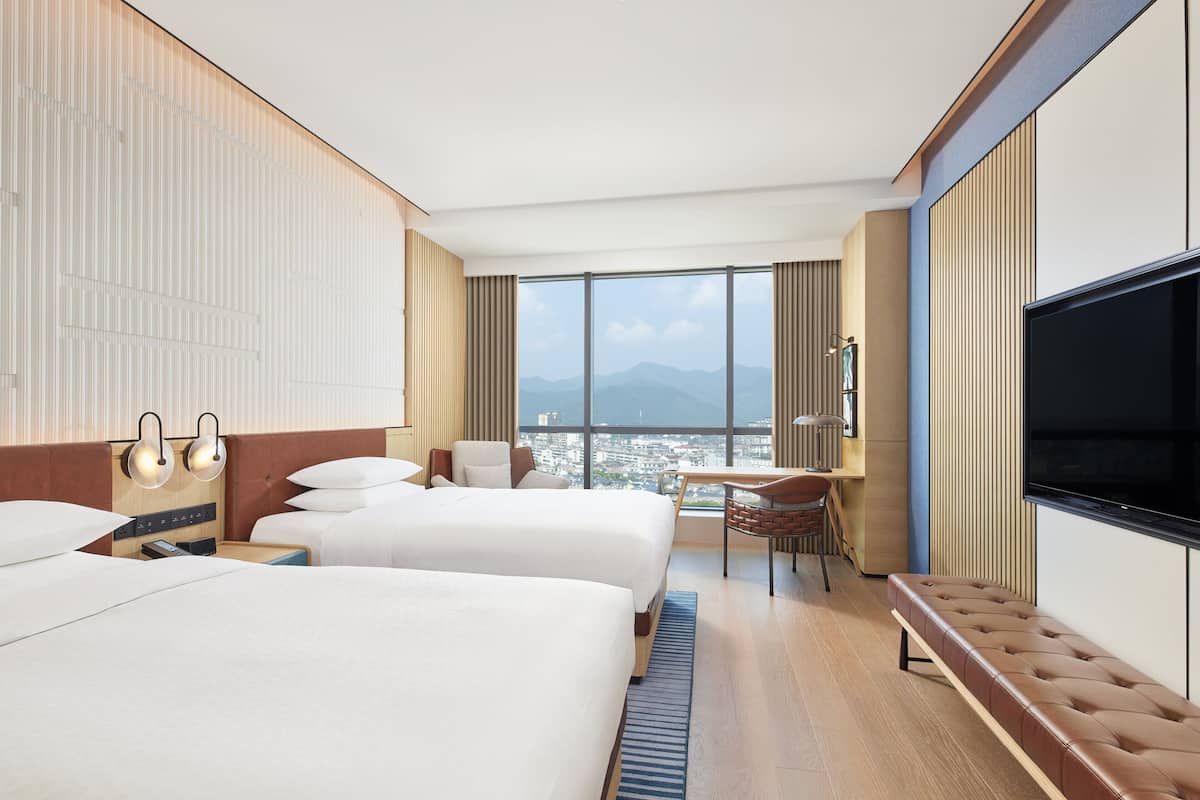 Habitación de hotel con dos camas, televisión, escritorio y vista a la ciudad.