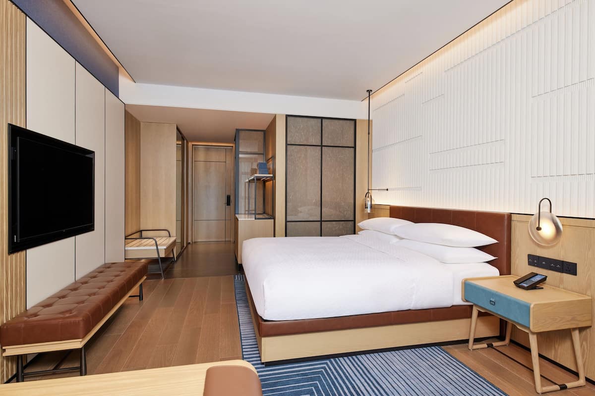 Una habitación de hotel moderna con una cama grande, un televisor de pantalla plana en la pared, una mesita de noche con lámpara y un banco.
