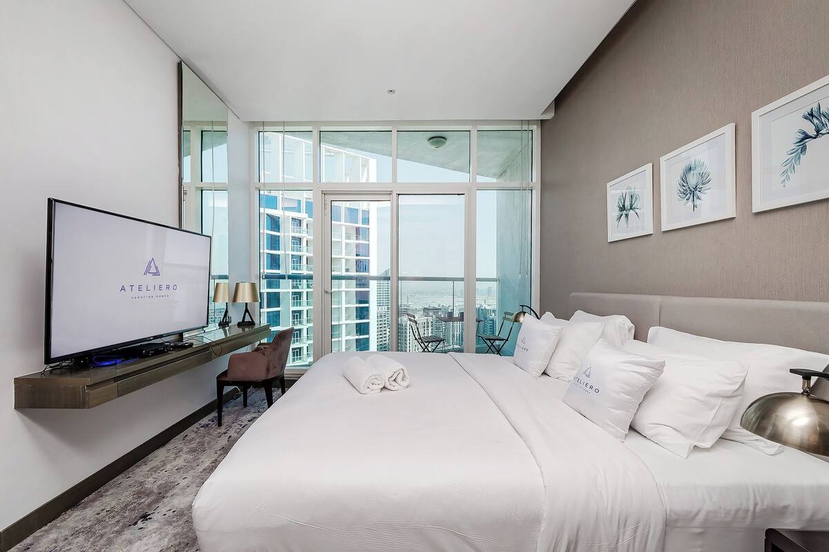 Une chambre d’hôtel moderne avec un grand lit, un téléviseur posé sur un meuble, un balcon offrant une vue sur la ville et des œuvres d’art encadrées accrochées au mur.