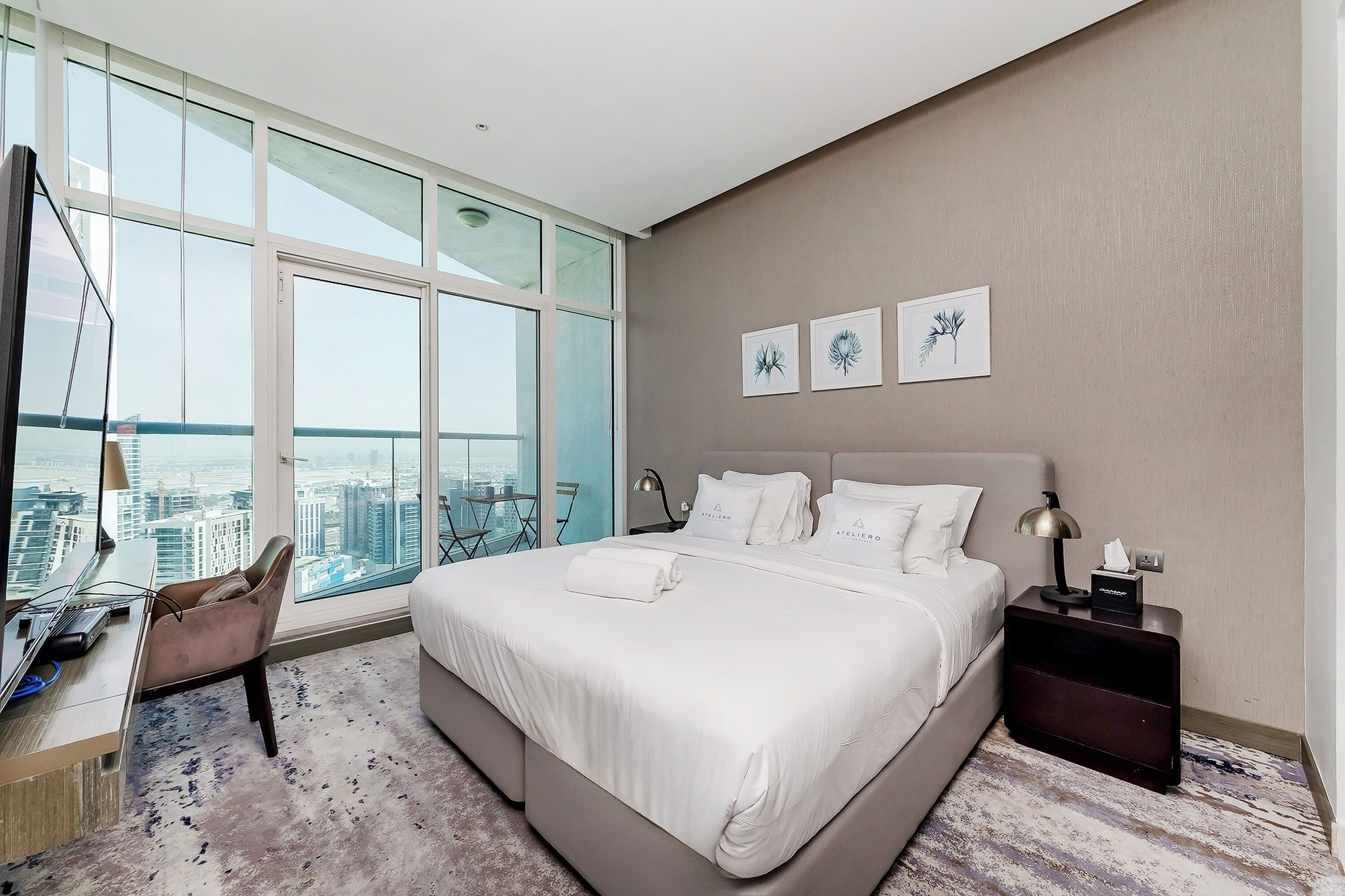 A modern hotel room with a large bed, a desk, and a view of the city through floor-to-ceiling windows.