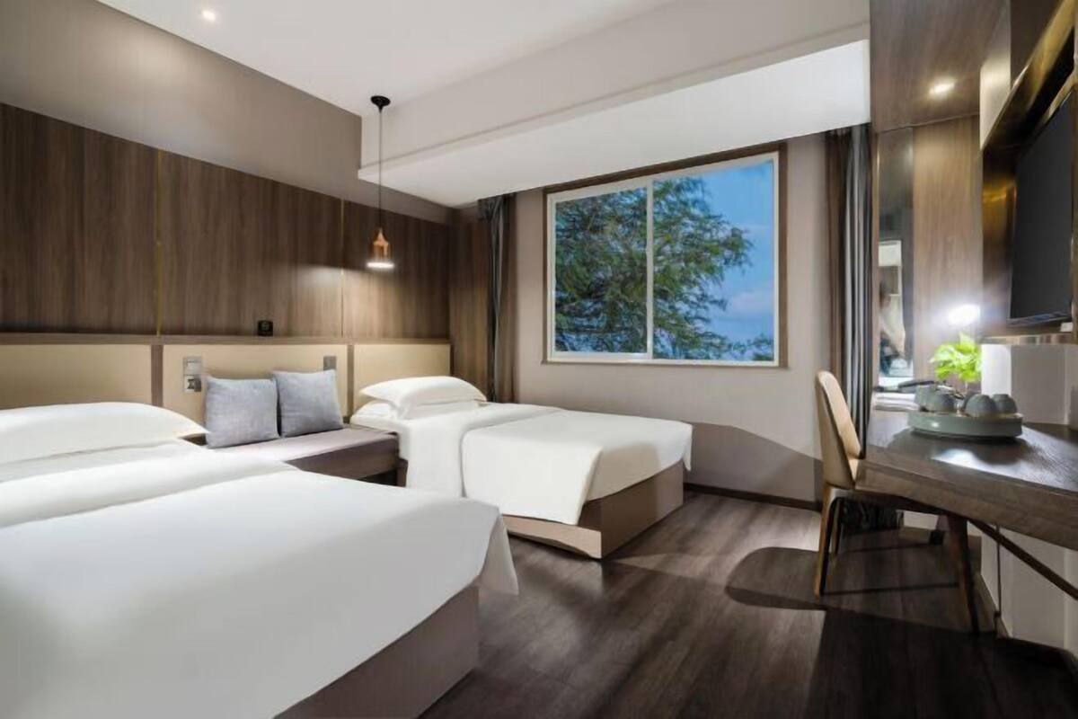 A modern hotel room with two beds, a desk, and a large window offering a view of trees.