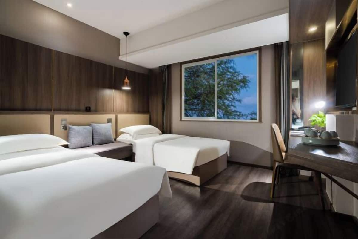 A hotel room with two beds, a desk, a chair, a window with a view, and a pendant light.