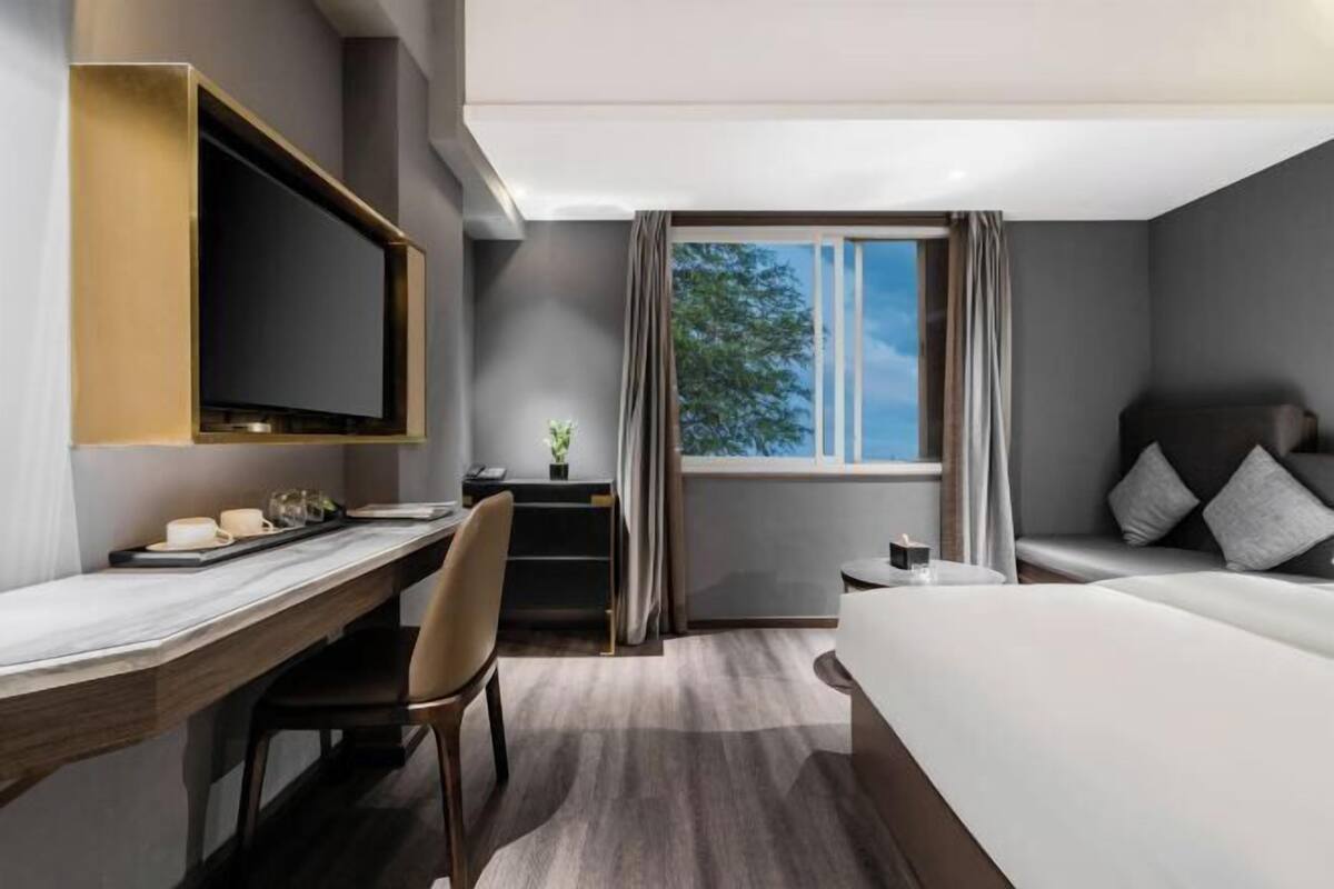 A hotel room with a bed, a desk, a television, a window with curtains, and a view of trees.
