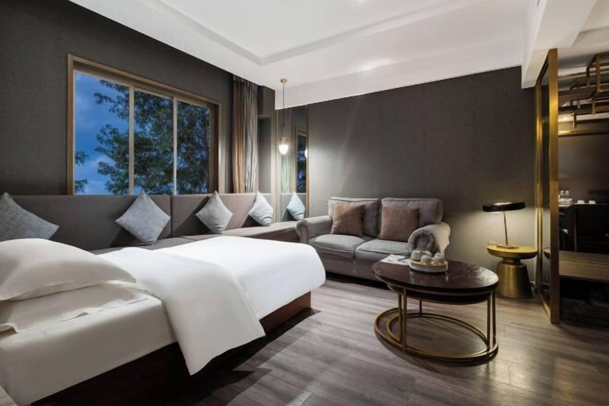A modern hotel room with a large bed, a sofa, a small table, and a view of trees through the window.