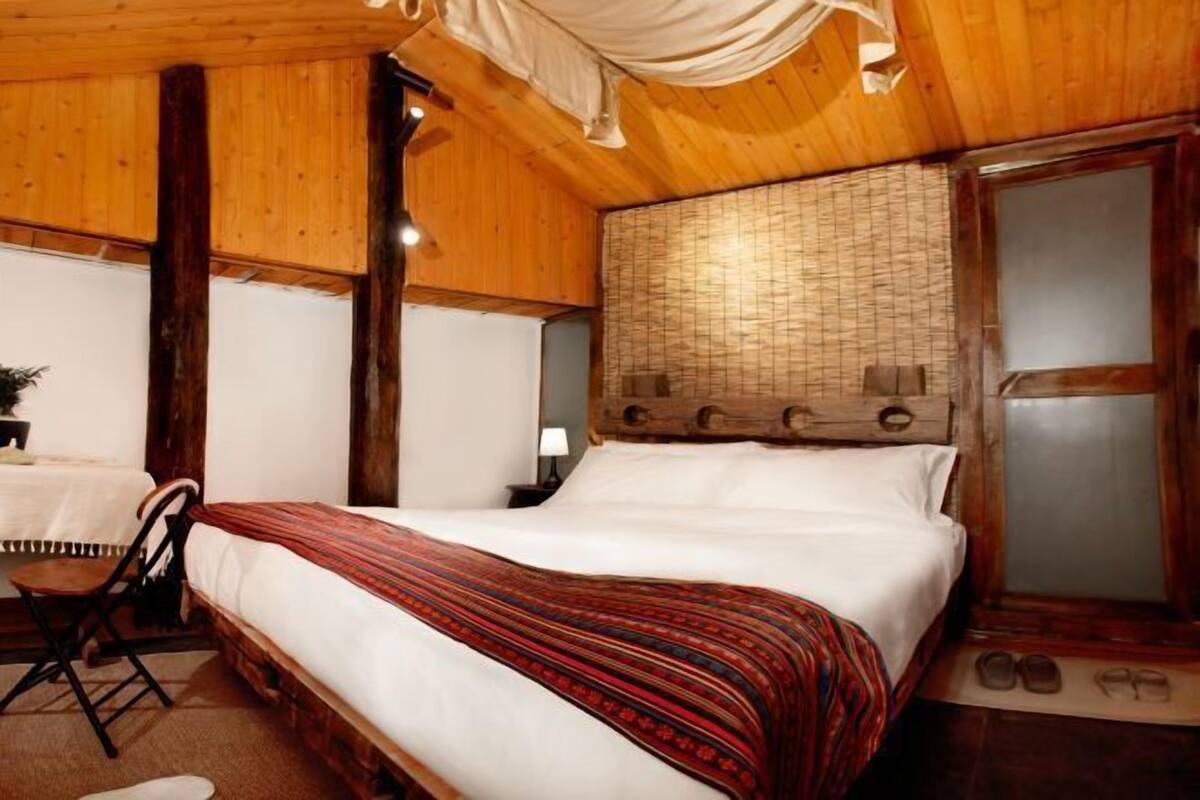 A wooden room with a bed, a chair, a nightstand, and a window with curtains.