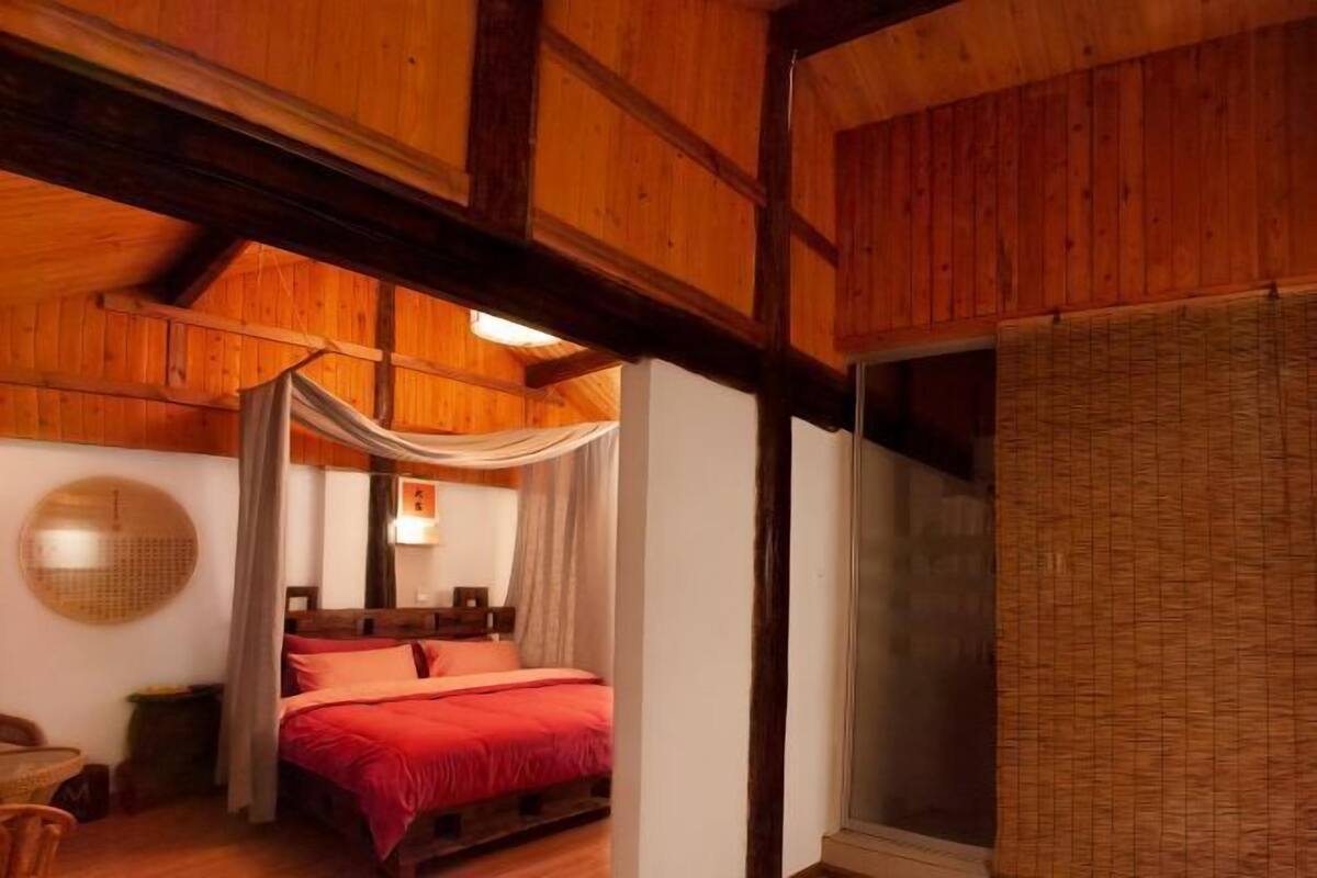 A wooden room with a bed, a round mirror, and a window with a woven blind.