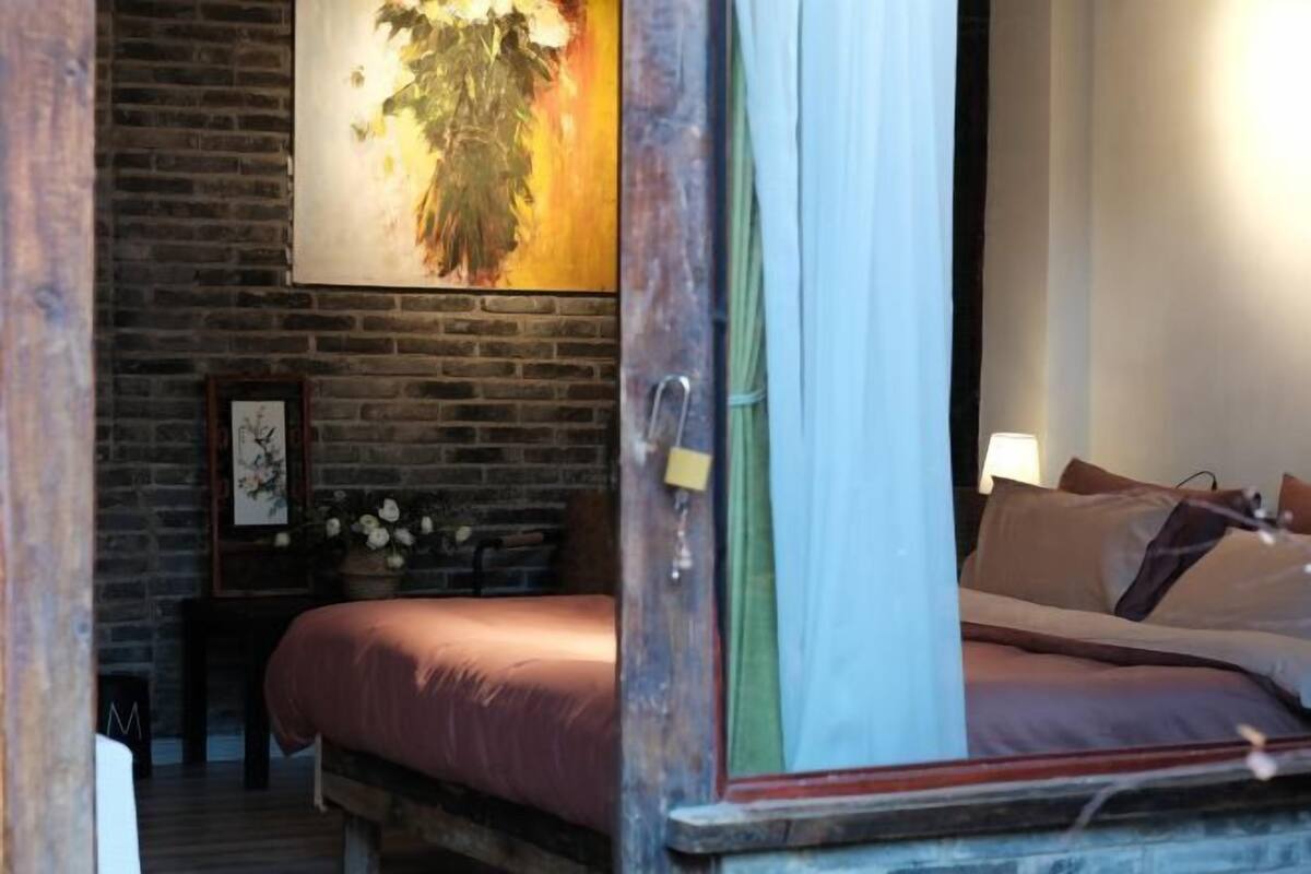 A room with a brick wall, a bed with a headboard, a bedside table with a lamp, and a framed picture on the wall.