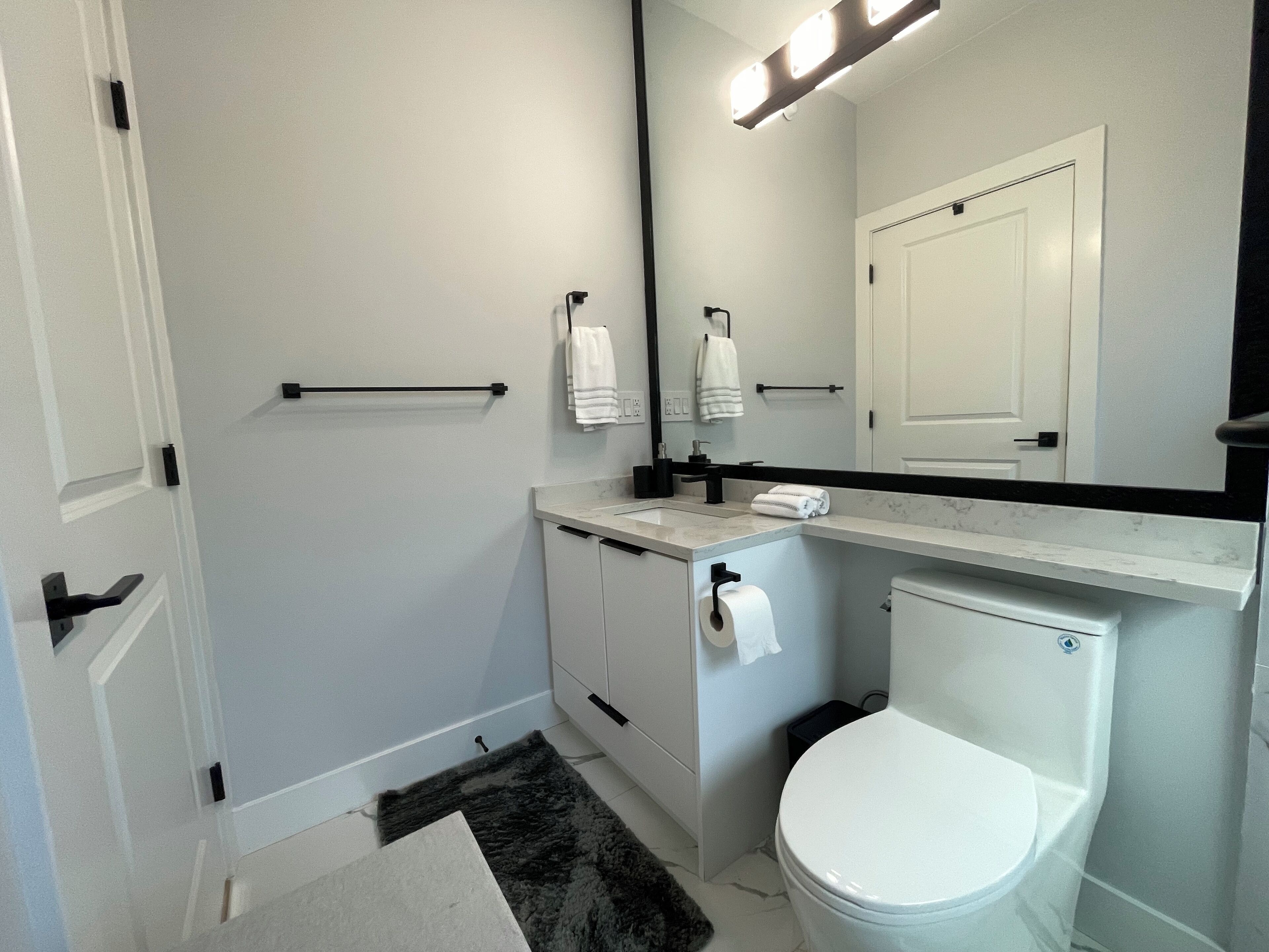 Ein modernes Badezimmer mit Toilette, Waschbecken mit schwarzem Wasserhahn, Handtuchhalter und einem Spiegel über dem Waschbecken.