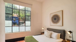 Ein Hotelzimmer mit großem Fenster, einem Bett mit weißer Bettwäsche, einer grünen Tagesdecke, zwei Nachttischen mit Lampen und einem gerahmten Kunstwerk an der Wand.