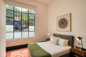 A hotel room with a large window, a bed with white bedding, a green throw, two bedside tables with lamps, and a framed artwork on the wall.