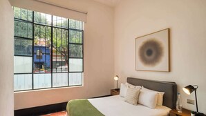 Habitación de hotel con un ventanal grande, una cama con ropa de cama blanca, un plaid verde, dos mesitas de noche con lámparas y una obra de arte enmarcada en la pared.