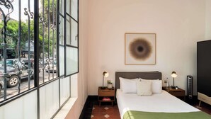 Habitación de hotel con un ventanal grande, una cama, dos mesitas de noche con lámparas, una obra de arte en la pared y un mueble negro para televisor.