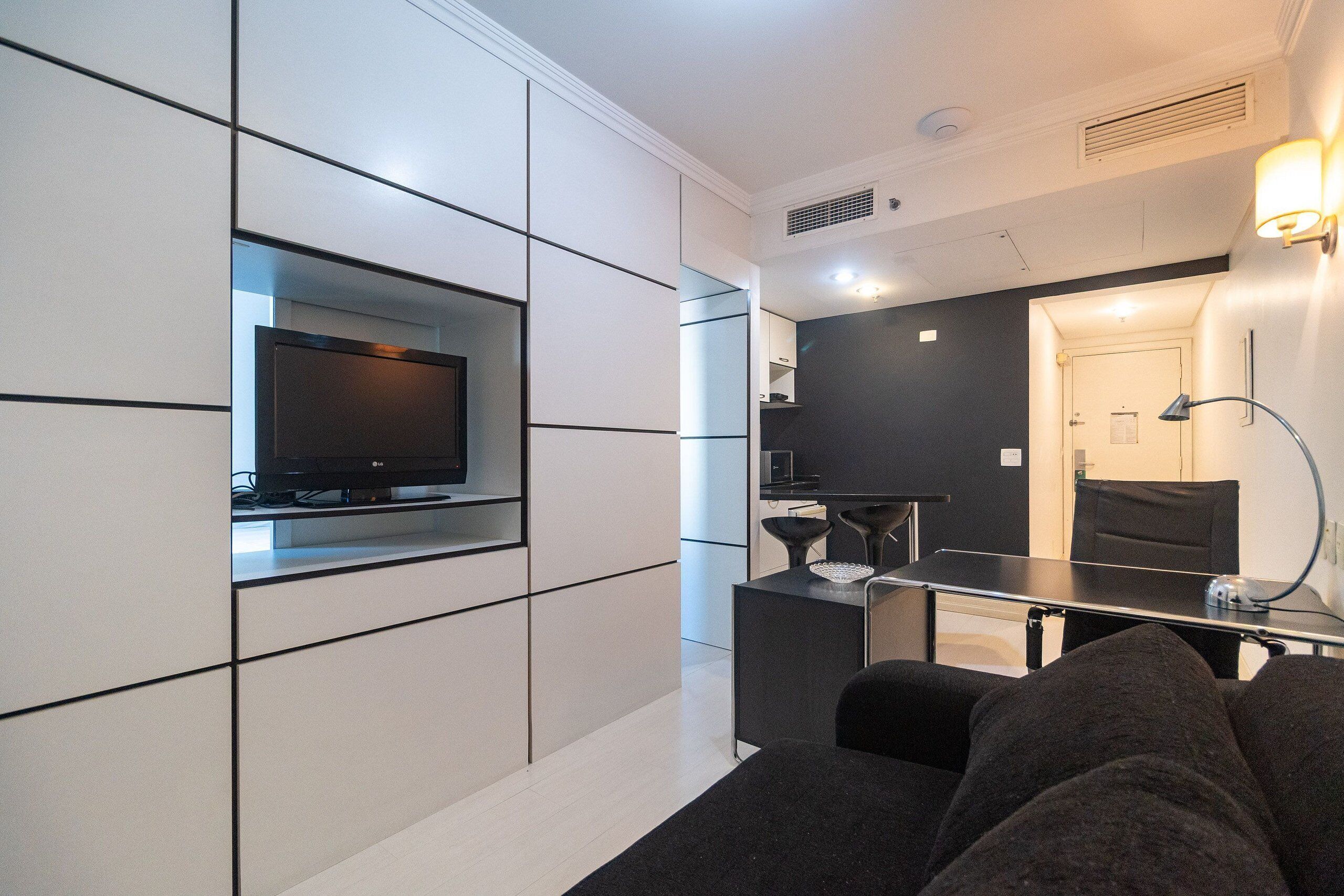 Une chambre d’hôtel moderne équipée d’un téléviseur à écran plat fixé au mur, d’une petite kitchenette, d’un canapé et d’un bureau avec une lampe.
