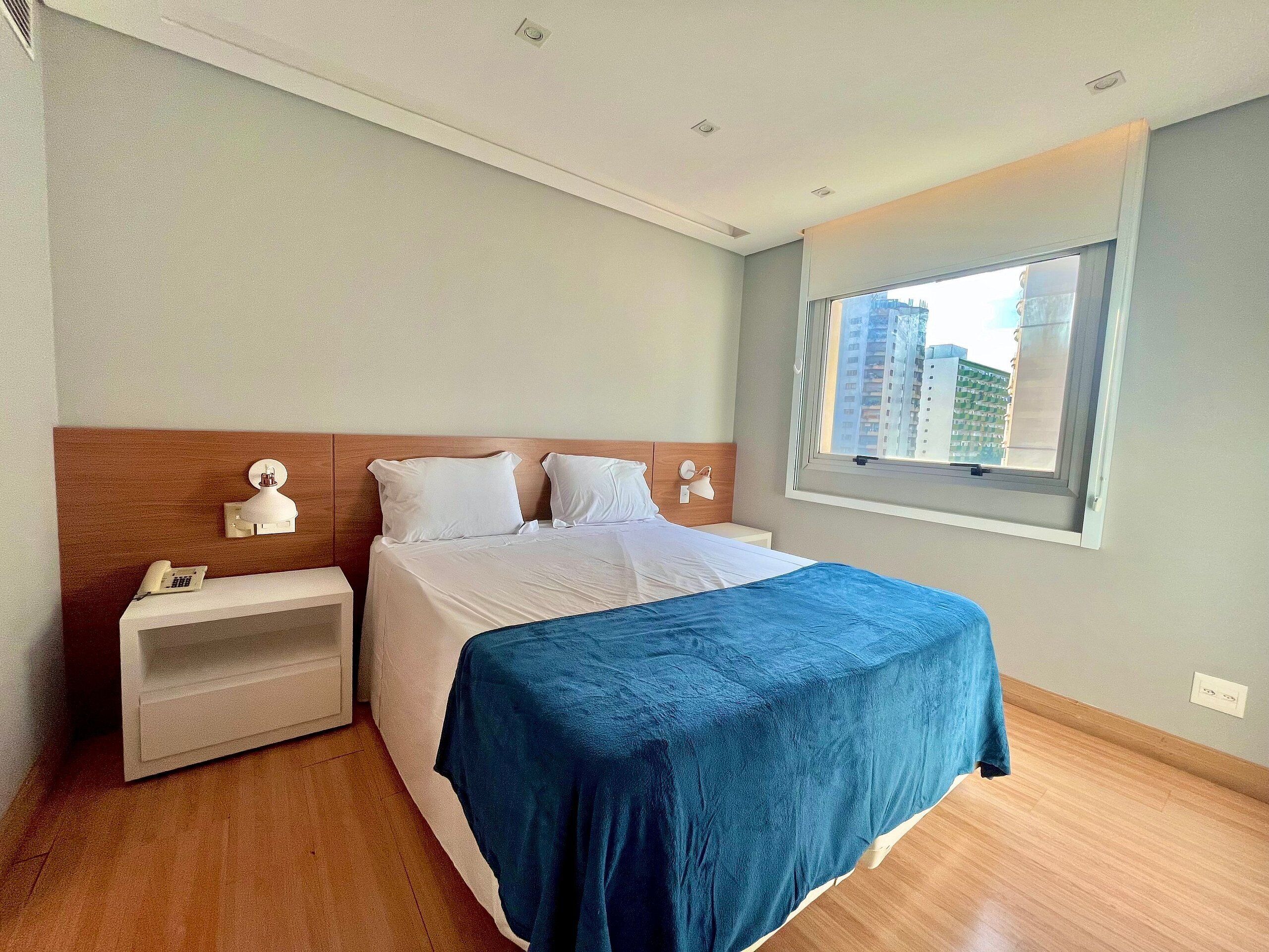 A hotel room with a bed, bedside tables, a window with a view of buildings, and a ceiling with recessed lighting.