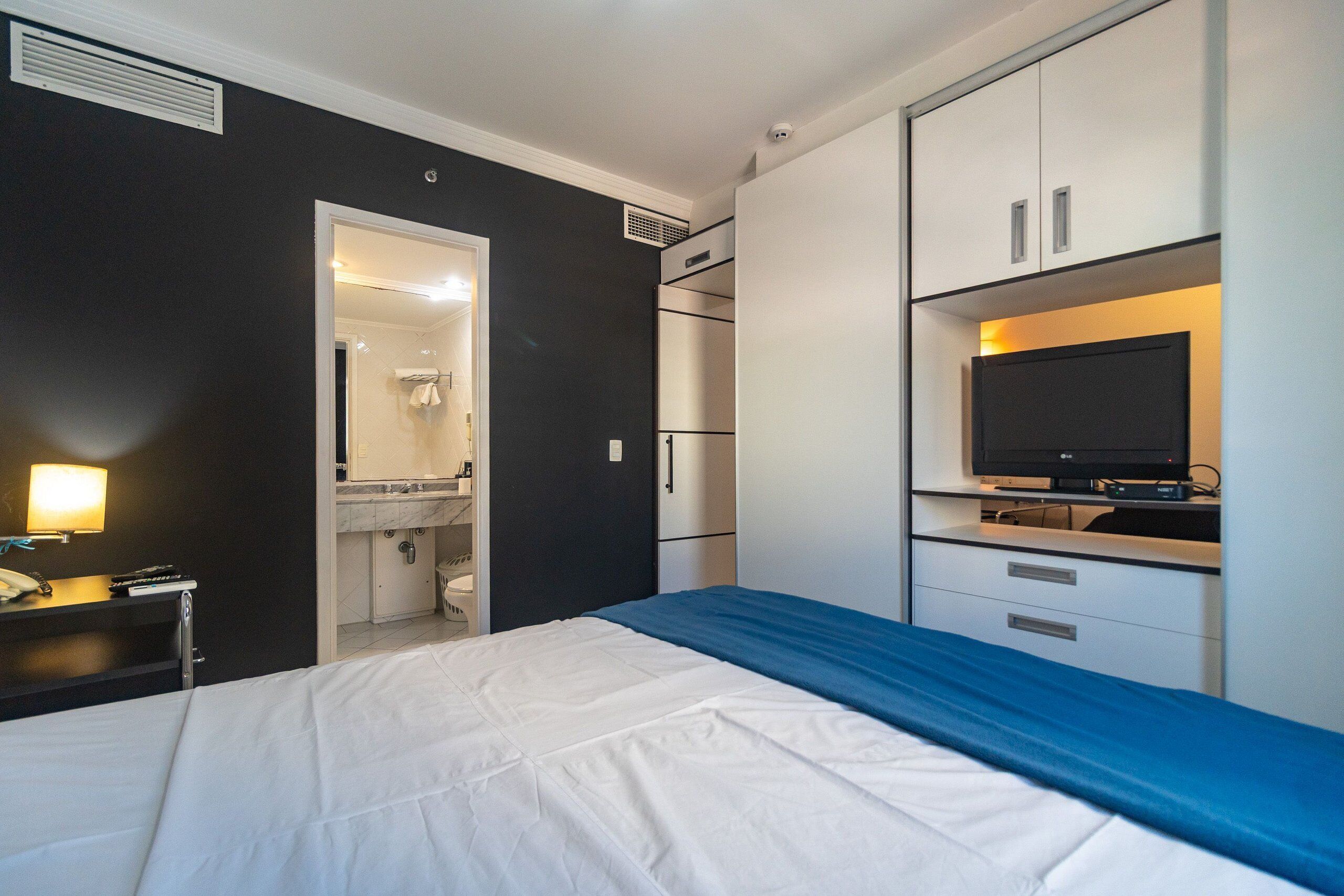 A hotel room with a bed, a desk with a lamp, a television, and a bathroom visible through an open door.