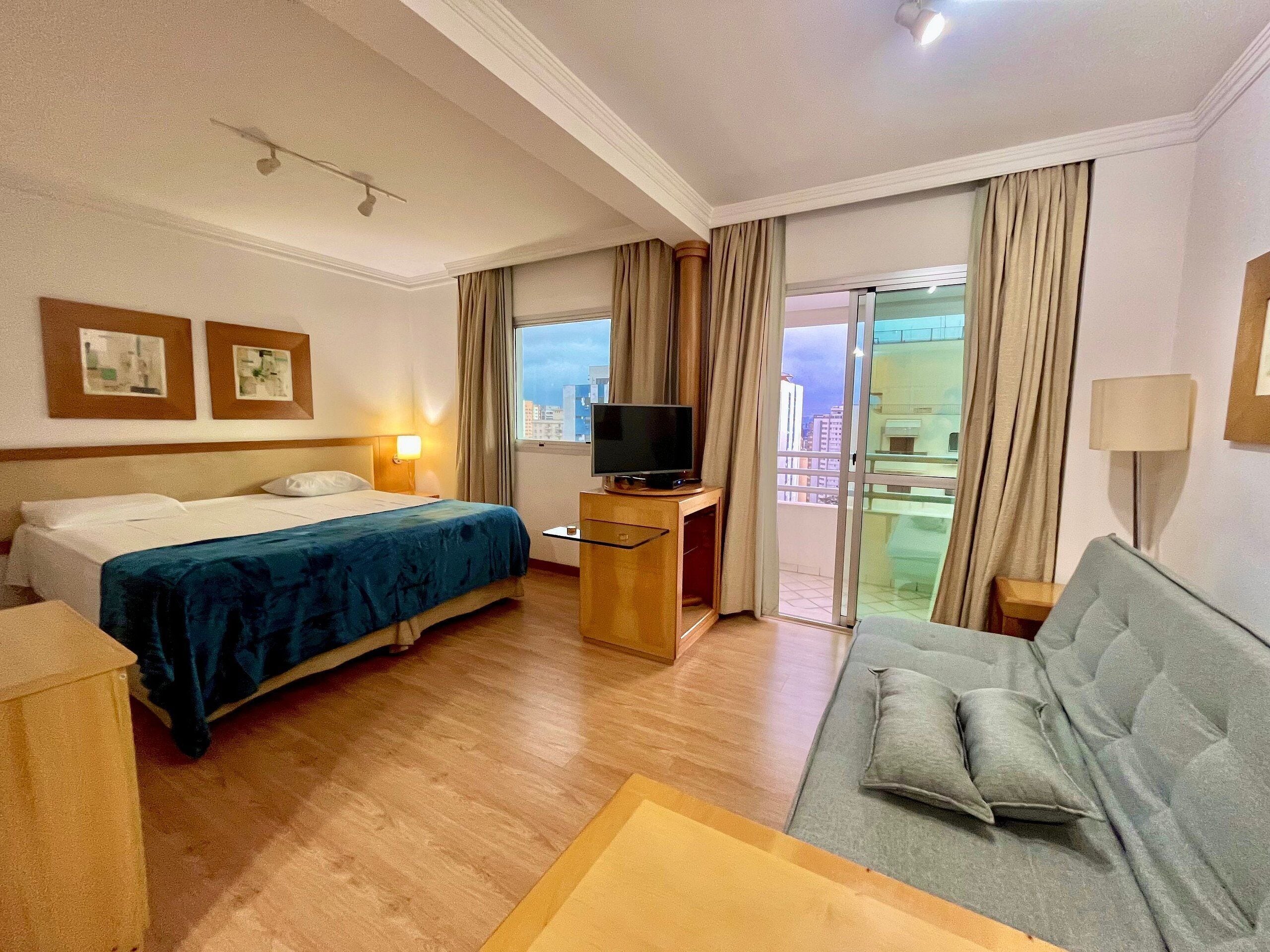 A hotel room with a bed, a sofa, a TV, a desk, and a balcony with curtains.