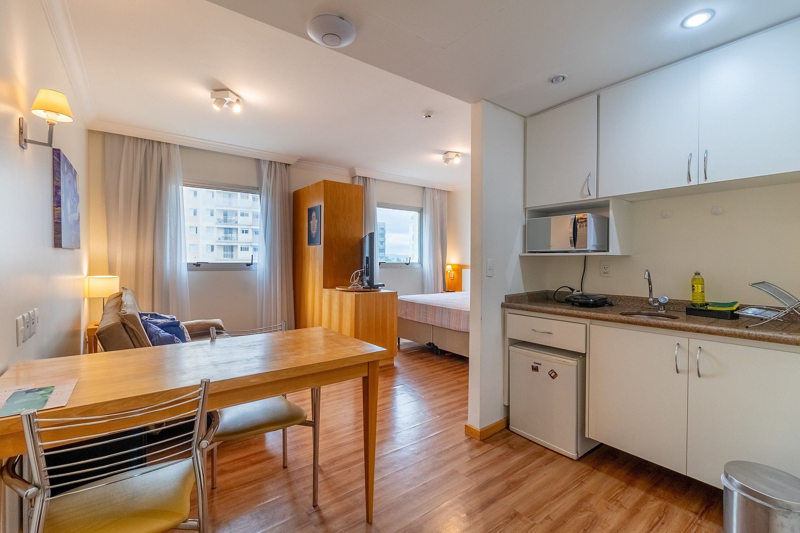 A compact hotel room with a wooden dining table, a small kitchenette, a bed, and a window with curtains.