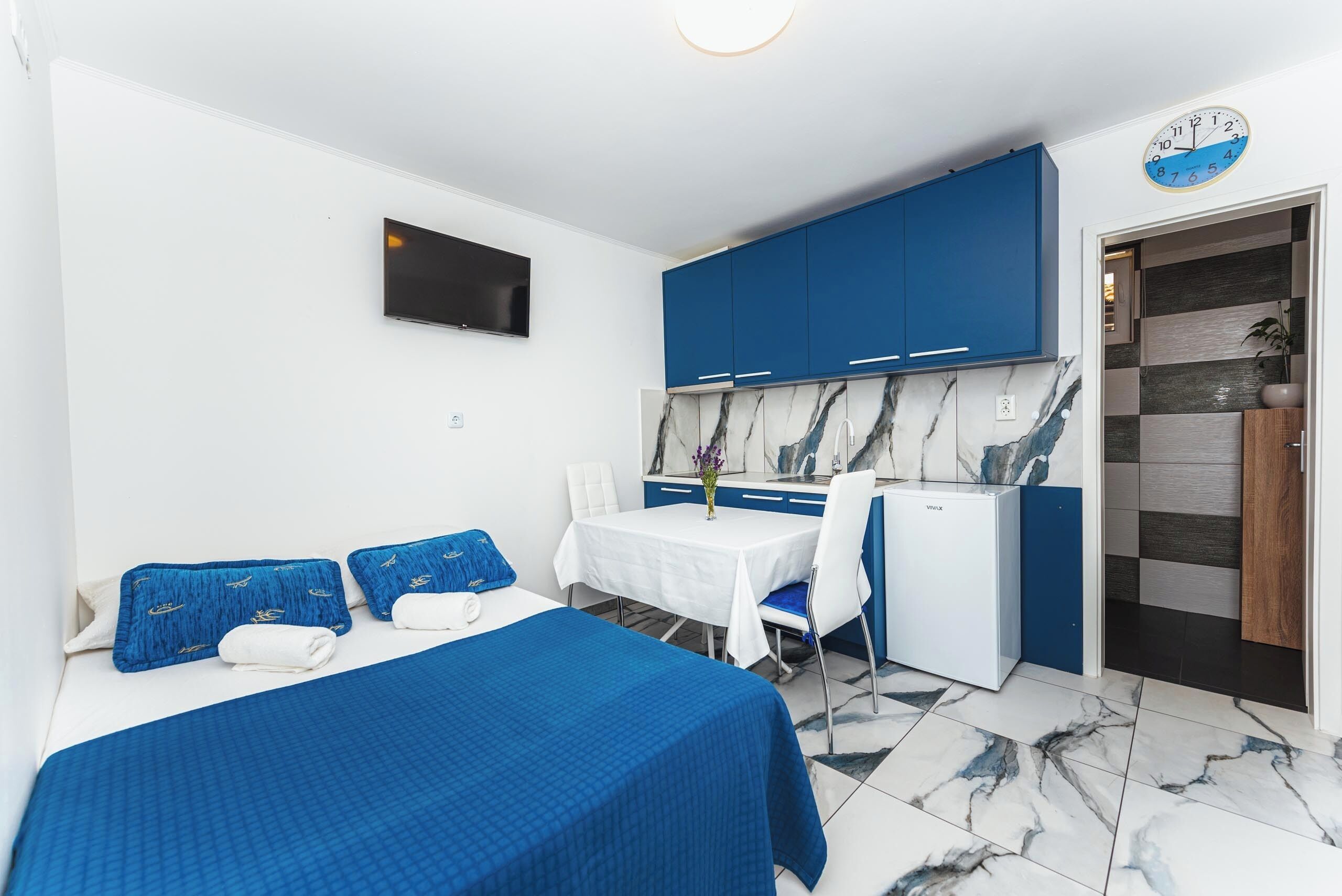Une chambre d’hôtel comprenant un lit, une petite table avec une chaise, une kitchenette et un téléviseur fixé au mur.